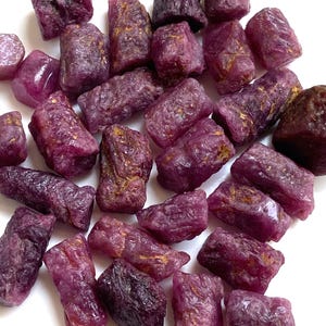 Może przedstawiać: Kolekcja surowych kamieni szlachetnych rubinu o różnych kształtach i rozmiarach. Kamienie wykazują głęboki czerwono-fioletowy odcień z naturalnymi teksturami i niektórymi obszarami o złoto-brązowym zabarwieniu. Kamienie szlachetne są rozrzucone na białej powierzchni.