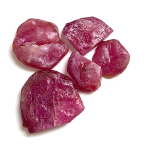 May include: A collection of raw, uncut ruby gemstones. The rubies display a deep red hue with variations in tone and texture. The stones are irregularly shaped, showcasing their natural, unpolished state. The image is set against a white background.