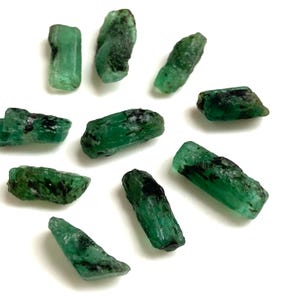 May include: A selection of raw emerald crystals, showcasing a rich green colour with dark markings. The gemstones are of varying sizes and shapes, set against a white backdrop. These natural emeralds are suitable for jewellery creation or as collector's items.
