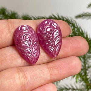 May include: Two oval-shaped, polished ruby gemstones. Each gemstone features a carved, symmetrical leaf design. The rubies are a deep pinkish-red color and are held in a hand, with a blurred green background.