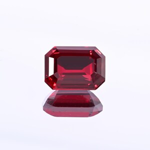 May include: A faceted, rectangular-cut, deep red gemstone. The gem is highly reflective, with a mirror image visible below. The stone's edges are precisely cut, showcasing its clarity and brilliance. The gem is set against a white background.
