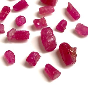 May include: A scattering of raw ruby gemstones, each with a deep red colour. The stones are of varying sizes and shapes, with a rough, natural texture. The rubies are displayed against a plain white background.
