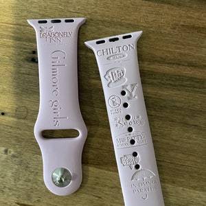 Peut inclure: Deux bracelets roses pour Apple Watch avec des motifs gravés. Le bracelet de gauche porte le texte "Gilmore Girls" et le texte "Dragonfly Inn" avec un logo de libellule. Le bracelet de droite porte le texte "Chilton Alumni" avec divers logos et textes, notamment "Lukes", "Snow", "Miss Patty's School of Ballet", "Doose's Market" et "In Omnia Paratus".
