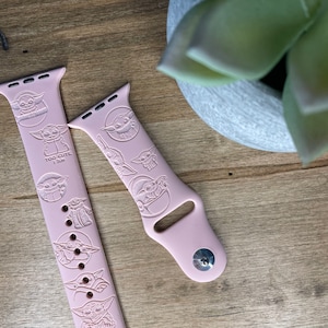 May include: Two pale pink Apple Watch straps featuring engraved images of a character. One strap has the text "TOO CUTE I AM". The straps are on a wooden surface, with a succulent plant in a grey pot in the background.