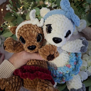 May include: Two handmade plush deer toys. One is brown with a red skirt and white antlers. The other is white with a blue bow and a blue floral skirt. Both have large black eyes and are made of soft, textured yarn.