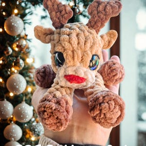 Könnte beinhalten: Ein handgefertigtes Rentierspielzeug mit braunem Fell, großen, irisierenden Augen und einem roten Mund. Der Text "Ruby The Reindeer" steht oben und "PDF Pattern Only" unten. Das Rentier wird in der Hand einer Person gehalten.