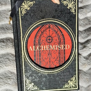 Puede incluir: Libro negro con adornos dorados y la palabra "ALCHEMISED" en la portada. La portada presenta un círculo rojo con un diseño de arco gótico. Se adjunta un marcador de cinta negra con un dije dorado.