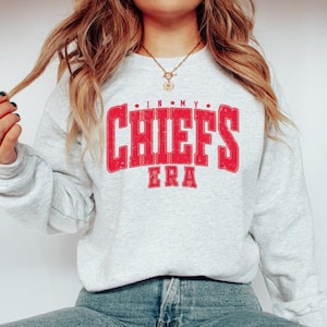 May include: A light gray sweatshirt with the text "IN MY CHIEFS ERA" in red, distressed lettering. The sweatshirt has long sleeves and a crew neck. The overall theme is sports apparel.