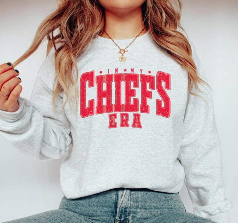 May include: A light gray sweatshirt with the text "IN MY CHIEFS ERA" in red, distressed lettering. The sweatshirt has long sleeves and a crew neck. The overall theme is sports apparel.