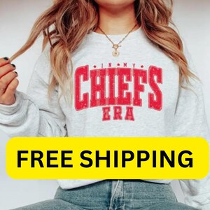 May include: A light gray sweatshirt with the text "IN MY CHIEFS ERA" in red. The sweatshirt has long sleeves and a crew neck. The image also includes a yellow banner with the text "FREE SHIPPING".