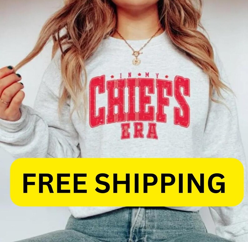 May include: A light gray sweatshirt with the text "IN MY CHIEFS ERA" in red. The sweatshirt has long sleeves and a crew neck. The image also includes a yellow banner with the text "FREE SHIPPING".