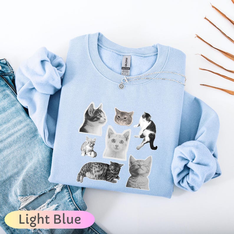 Op de afbeelding: Lichtblauw sweatshirt met een collage van kattengezichten in verschillende poses en kleuren. Het sweatshirt heeft lange mouwen en een ronde hals. Een zilveren ketting met een kleine hanger is zichtbaar. De tekst "Light Blue" staat onderaan.