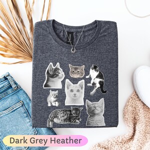 Op de afbeelding: Donkergrijze heather t-shirt met illustraties van katten in verschillende poses en kleuren. De shirt heeft een ronde hals en een zilveren ketting met een zonnehanger. De tekst "Dark Grey Heather" staat onderaan.