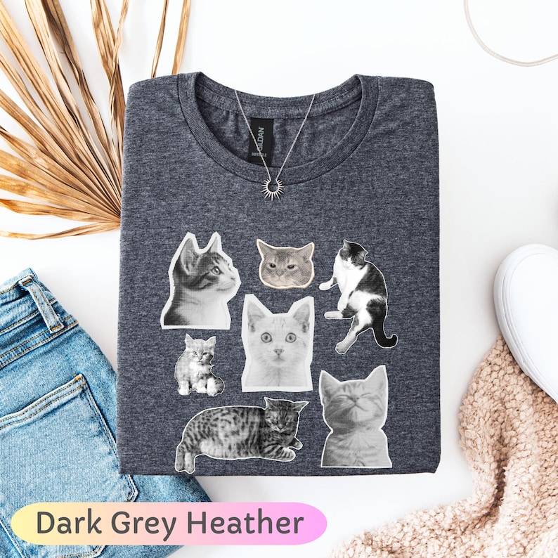 Op de afbeelding: Donkergrijze heather t-shirt met illustraties van katten in verschillende poses en kleuren. De shirt heeft een ronde hals en een zilveren ketting met een zonnehanger. De tekst "Dark Grey Heather" staat onderaan.