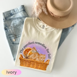 May include: Ivory-colored t-shirt with a graphic of a cat in a basket, an orange slice, and the text "The purrfect remedies for fall." The shirt is paired with light blue jeans, a beige hat, and a necklace.