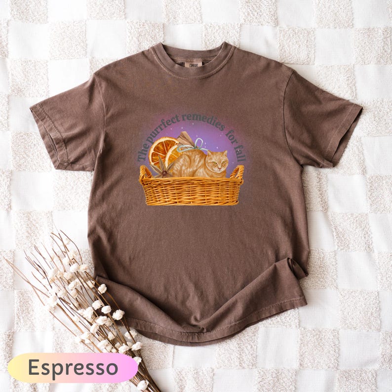 May include: Espresso-colored t-shirt featuring a graphic of a ginger cat in a wicker basket with dried oranges and the text "The purrfect remedies for fall." The shirt is a comfortable, casual style.