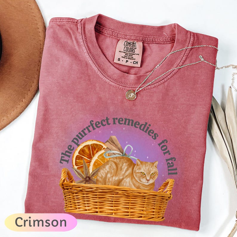 May include: Crimson-colored t-shirt with a graphic of a cat in a basket, surrounded by orange slices and spices. The text reads "The purrfect remedies for fall". A silver necklace is draped across the shirt.
