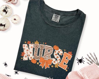 May include: Dark green t-shirt with the word "NURSE" in a fall-themed design. The letters feature patterns like leopard print, stripes, and polka dots, with pumpkins and leaves. The shirt is surrounded by Halloween decorations.