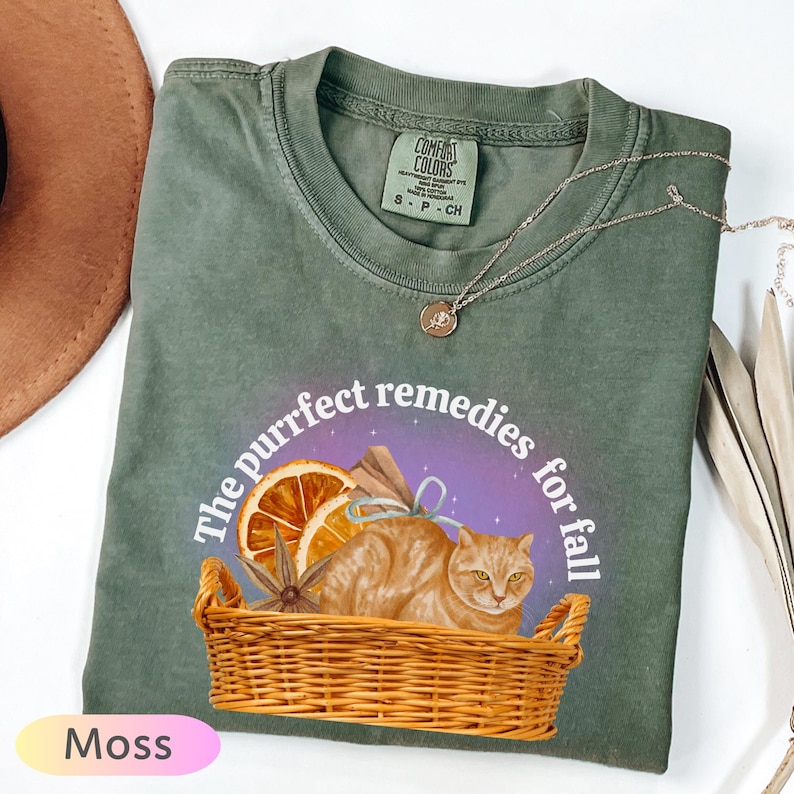 May include: A moss green t-shirt with a graphic design featuring a cat in a basket, orange slices, and the text "The purrfect remedies for fall." The shirt also has a Comfort Colors tag and a small necklace.