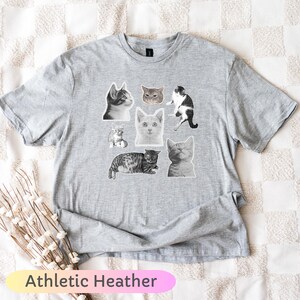 Op de afbeelding: Athletic Heather grijs T-shirt met een collage van kattenillustraties. De katten worden afgebeeld in verschillende poses en patronen, waaronder zwart-wit, grijs en tabby. De tekst "Athletic Heather" staat onderaan.