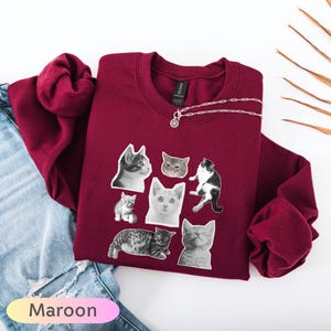 Op de afbeelding: Bordeaux sweatshirt met een ronde hals en lange mouwen, versierd met een collage van zwart-wit kattenafbeeldingen. Een zilveren ketting is eroverheen gedrapeerd. Het woord "Maroon" wordt in een verlooptekst linksonder in de afbeelding weergegeven.