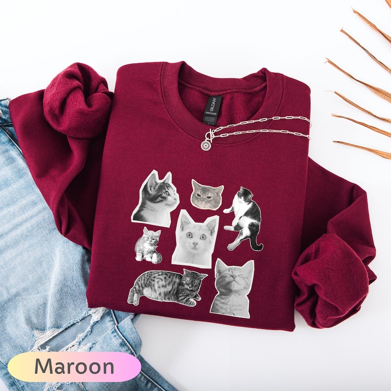 Op de afbeelding: Bordeaux sweatshirt met een ronde hals en lange mouwen, versierd met een collage van zwart-wit kattenafbeeldingen. Een zilveren ketting is eroverheen gedrapeerd. Het woord "Maroon" wordt in een verlooptekst linksonder in de afbeelding weergegeven.