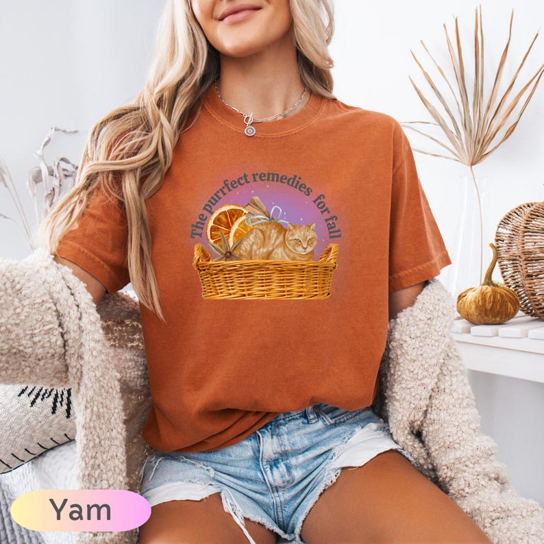 May include: Orange-colored t-shirt with a graphic of a cat in a basket, orange slices, and text that reads "The purrfect remedies for fall." The shirt is made of a soft material.