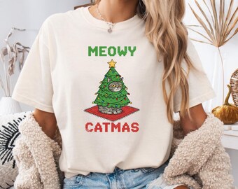 Puede incluir: Camiseta color marfil con las palabras "MEOWY CATMAS" en texto pixelado verde y rojo. El gráfico presenta un gato disfrazado de árbol de Navidad sobre una alfombra roja. La camiseta es de corte holgado.