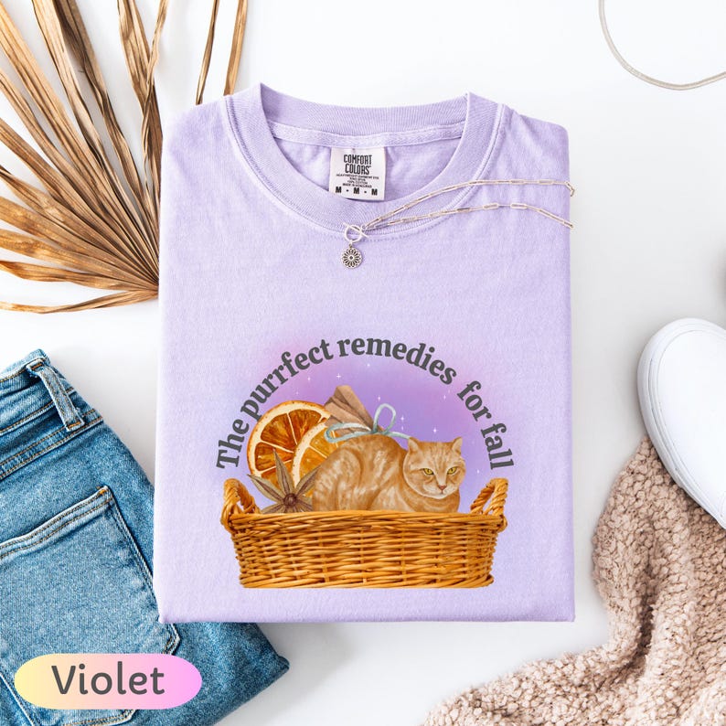 May include: Violet Comfort Colors t-shirt with a graphic of a cat in a basket with the text "The purrfect remedies for fall." The shirt is folded with a silver necklace on top. A pair of blue jeans and a beige sweater are visible.