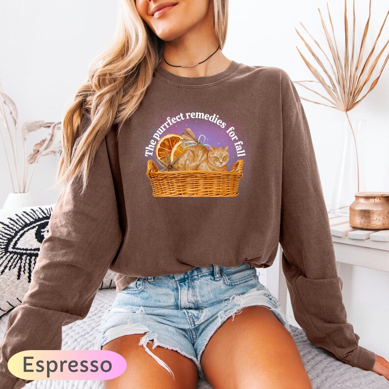 May include: Espresso-colored sweatshirt featuring a graphic of a cat in a basket with orange slices. The text reads "The purrfect remedies for fall." The sweatshirt is a long-sleeved crew neck.