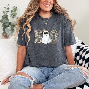 May include: Dark gray t-shirt featuring the word "BOO" with a ghost graphic. The letters are in a camouflage pattern, and the ghost is wearing a camouflage hat. The shirt is a comfortable, casual style.