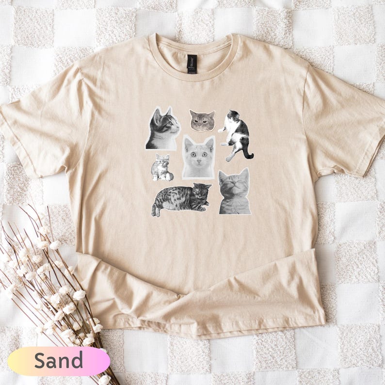 Op de afbeelding: Zandkleurig T-shirt met een verzameling zwart-wit kattenillustraties. De katten worden afgebeeld in verschillende poses en uitdrukkingen, wat een speels ontwerp cre&euml;ert. Het shirt is gemaakt van een zacht materiaal.