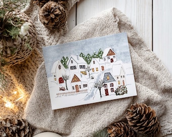 May include: A watercolor greeting card depicting a snow-covered village with white houses, red doors, and lit windows. The card is surrounded by pine cones, a beige knitted blanket, and a white ceramic mug, evoking a cozy winter theme.