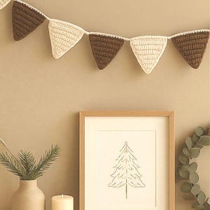 May include: A decorative crochet garland featuring alternating brown and cream-coloured triangular flags. A framed tree sketch, a vase with greenery, and a candle on a wooden coaster create a cosy, rustic aesthetic.