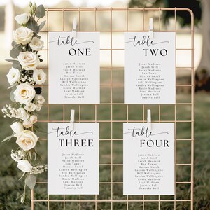 May include: A copper-colored grid holds four white cards with guest seating information. Each card is labeled "table" followed by a number and includes a list of names. White flowers and greenery decorate the left side, adding a touch of elegance.