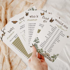 Op de afbeelding: Een hand houdt een stapel babyshower-spelkaarten vast. De kaarten bevatten botanische illustraties van vlinders en bloemen. Tekst op de kaarten is onder meer "Baby Name", "Who Knows Mom" en "Would She Rather?" De kaarten hebben een neutraal kleurenpalet.