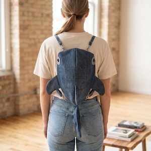 May include: A denim shark-shaped backpack with adjustable straps. The backpack features a blue color, with fins and a tail. The person is wearing a light-colored t-shirt and blue jeans.