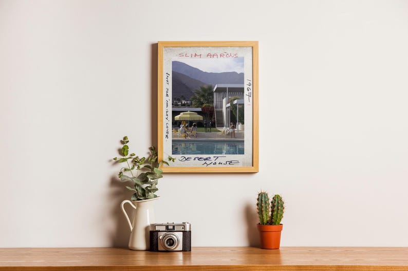 May include: A framed print of a photograph by Slim Aarons, titled "Desert House" with the year 1967. The image depicts a mid-century modern home with a pool, people, and a mountain backdrop. A vintage camera, a white pitcher with greenery, and a cactus in a terracotta pot sit below.