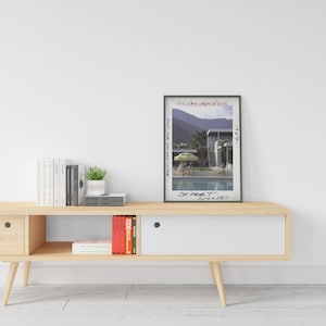 May include: A framed print of a photograph titled "Desert House" by Slim Aarons, 1967. The image depicts a mid-century modern home with a pool, people, and a yellow umbrella. The print is displayed on a light wood console table.