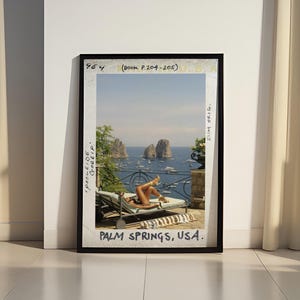 May include: Framed photograph featuring a sunny scene with a person relaxing on a lounge chair overlooking a blue sea with boats and islands. The frame includes handwritten text and the words "PALM SPRINGS, USA."