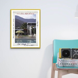 May include: A framed print with a yellow border featuring a photograph of a pool and a mid-century modern house. The print is titled "Desert House" by Slim Aarons. A yellow and black radio sits on a stack of magazines on a teal chair.