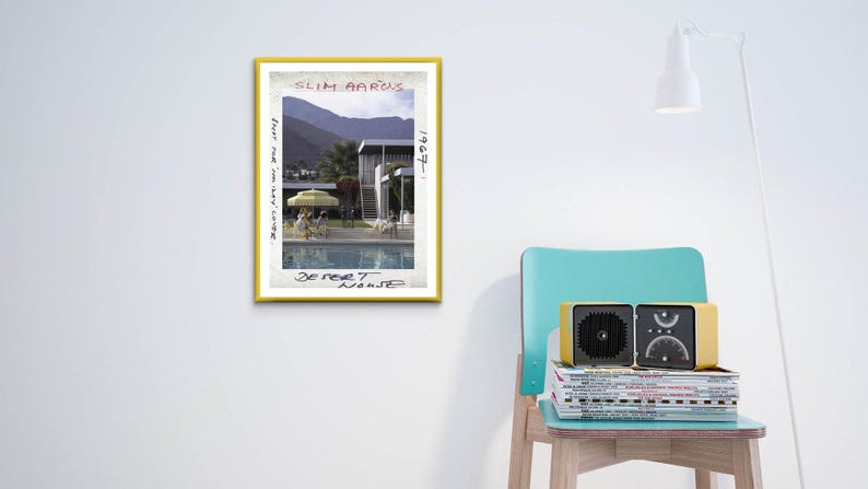 May include: A framed print with a yellow border featuring a photograph of a pool and a mid-century modern house. The print is titled "Desert House" by Slim Aarons. A yellow and black radio sits on a stack of magazines on a teal chair.