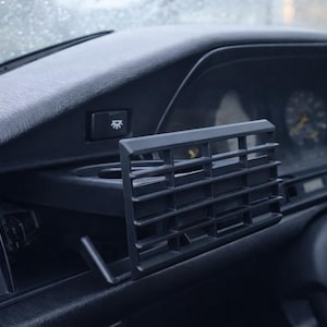 以下が含まれることがあります： 長方形の通気口、カップホルダー、ゲージ付きダッシュボードを備えた黒い車のダッシュボードのクローズアップ。 ダッシュボードはマット仕上げで、いくつかのボタンがあります。 画像はわずかに角度の付いた視点から撮影されています。
