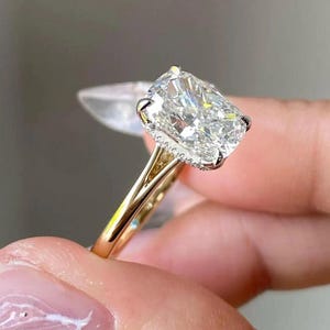 May include: A gold ring featuring a large, rectangular cushion-cut diamond set in a four-prong setting. The band is polished yellow gold, and the center stone displays multiple facets that reflect light, creating a brilliant, clear appearance.