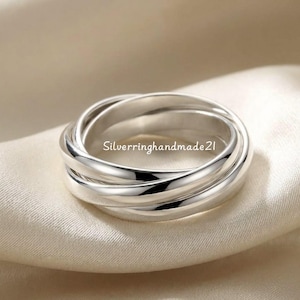 Op de afbeelding: Een zilveren ring met drie in elkaar grijpende banden. De banden zijn glad en glanzend en reflecteren licht. De ring wordt gepresenteerd op een zachte, neutraal gekleurde stof. De tekst "Silverringhandmade21" is zichtbaar op de ring.