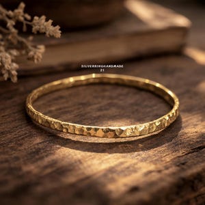 May include: A gold-colored bangle bracelet with a textured surface. The bracelet is circular and appears to be made of metal. The text "SILVERRINGHANDMADE 21" is visible in the image. The bracelet is displayed on a wooden surface.