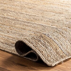 May include: A rectangular area rug crafted from natural tan and beige woven fibers. The rug features a thick, braided texture with a visible weave pattern. One corner of the rug is folded over, revealing the underside and the dense, layered construction.
