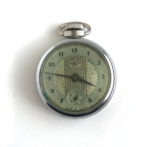 May include: A silver-toned Westclox pocket watch with a round face, black Arabic numerals, and a small sub-dial for seconds. The dial features a pale green speckled background with vertical lines and the text Westclox Dax Made in U.S.A.
