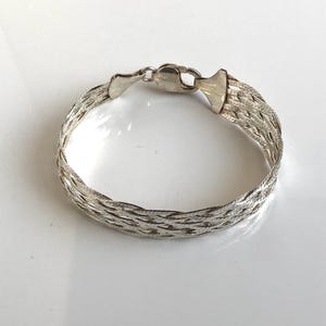 May include: A silver-toned bracelet featuring a multi-strand braided design with a polished metallic finish. The piece includes a secure lobster claw clasp mechanism that connects the woven metal segments into a continuous circular loop.