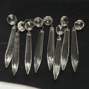 May include: Clear crystal chandelier prisms with faceted edges and octagonal top beads. The prisms are elongated, teardrop-shaped, and connected by thin wire. The crystals reflect light, creating a sparkling effect.